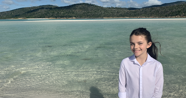 Lily wearing Qantas Choir white shirt and black pants in front of crystal clear blue water at Whitehaven beach