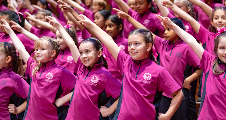 Four rows of choristers in magenta pink AGC t-shirt smiling with one hand on hip and the other diagonal to the sky.