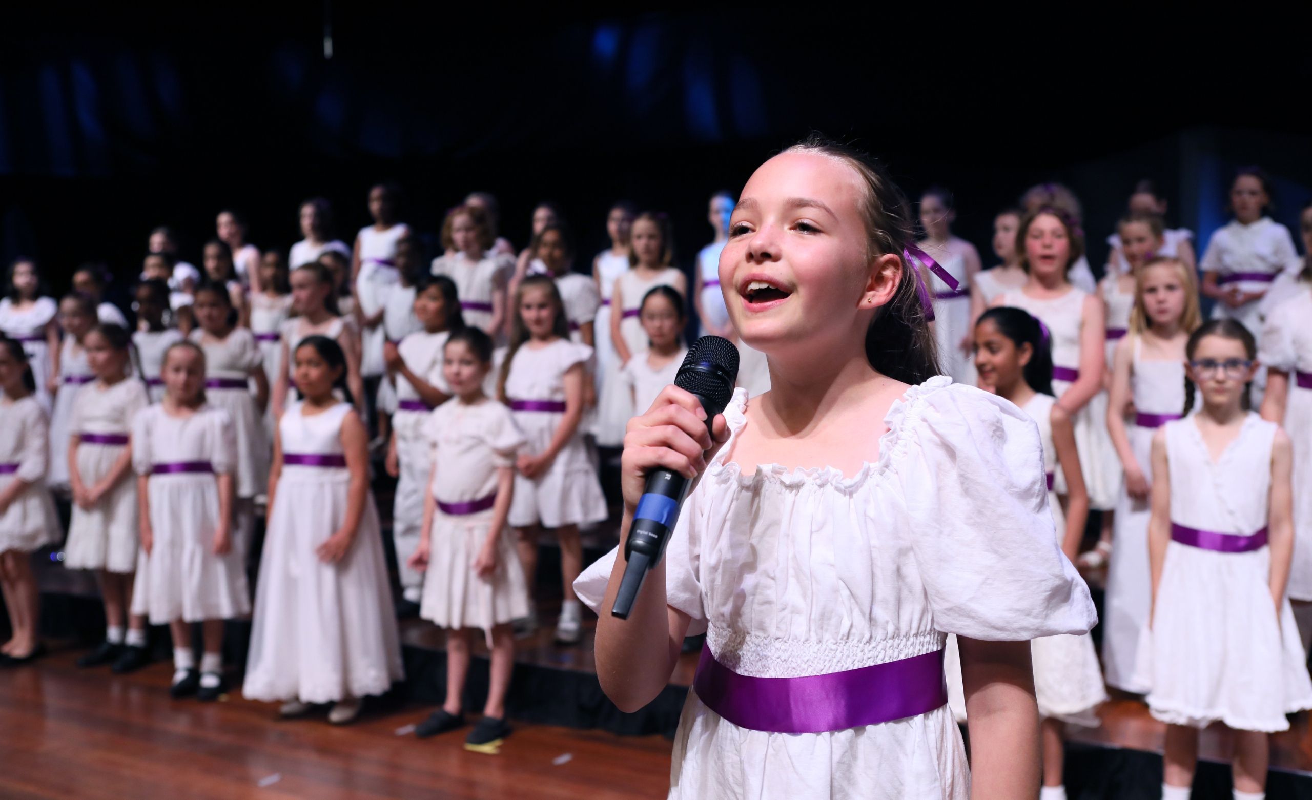 junior girls choir performing in on stage in white and purple dresses