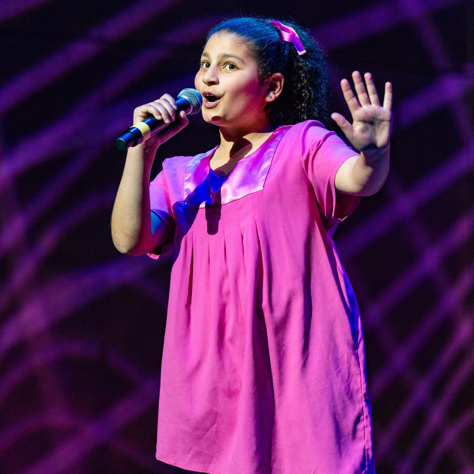 Training level chorister sings into a microphone with her arm out on stage